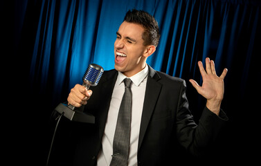 Man dressed in a business suit singing or yelling into a microphone on a  blue curtain background