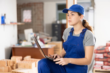 Young female designer in blue overalls working with laptop in apartment under renovation, planning remodeling and interior finishing