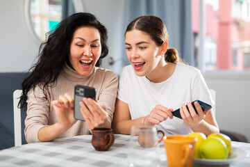 Two young adult smiling women using mobile phones at home, messaging or checking social media