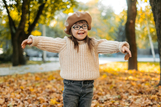 Happy cute brown-haired woman with Down's syndrome in a knitted sweater and stylish glasses throws bright foliage into the sky and laughs, the child enjoys time in a warm autumn park, happy childhood