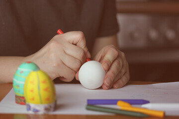 Fototapeta na wymiar Coloring Easter eggs with red wax crayon in home kitchen. White Easter egg in women's hands before dyeing colors. Easter egg festival. Artistic work, handmade DIY. People preparing for Easter in April