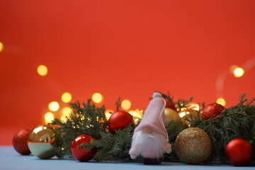 A small gnome stands in a festive atmosphere against the background of the branches of a Christmas tree with New Year's lights, and Christmas balls lie around