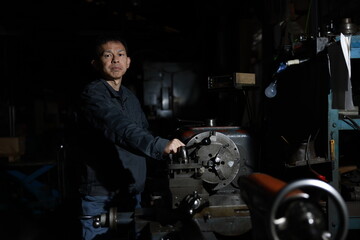 A craftsman poses in gray work clothes in front of a lathe at a local factory. Conceptual images of the essence of manufacturing, technical succession, and the challenge of high-precision machining.