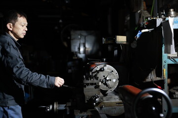 Obraz na płótnie Canvas A craftsman poses in gray work clothes in front of a lathe at a local factory. Conceptual images of the essence of manufacturing, technical succession, and the challenge of high-precision machining.