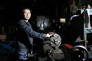 A craftsman poses in gray work clothes in front of a lathe at a local factory. Conceptual images of the essence of manufacturing, technical succession, and the challenge of high-precision machining.