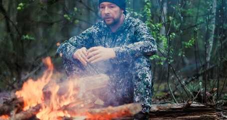 Man with cup of tea the fire in the forest. Human looks at the bonfire and drinks tea on the nature in the summer. Alone traveler sits and rests by the fire with a cup of coffee at dusk.