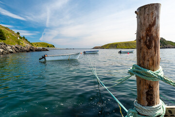 Fototapeta premium A large wood mooring with a green rope tied to the log. The cove has calm green colored water with three small open boats and two small grass covered islands. The sky is cloudy blue. 