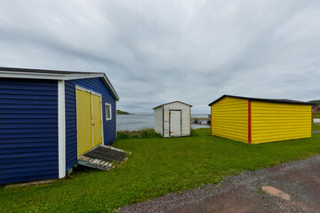 Multiple colorful wooden beach huts on vibrant green grass. The small buildings are yellow, blue, red, and white colored. The blue shed has a vibrant double yellow door with a ramp up the door.