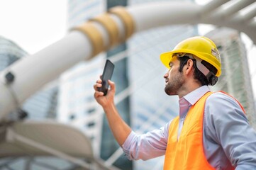 Caucasian man engineers use a smartphone for talking, wearing an orange vest and a big hard hat, and the other hand holding the white floor plan in the site work of the center city.