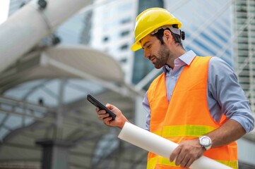 Caucasian man engineers use a smartphone for talking, wearing an orange vest and a big hard hat, and the other hand holding the white floor plan in the site work of the center city.