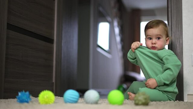 Beautiful toddler sitting at the doorway on the floor pulling his shirt looking at camera. Baby stands on all four and crawls away. Little balls at foreground.