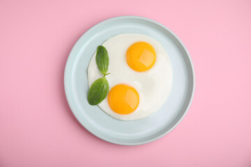 Tasty fried eggs with basil in plate on pink background, top view