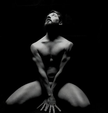 nude artistic male model looking up into light in pose accenting muscular definition with black background