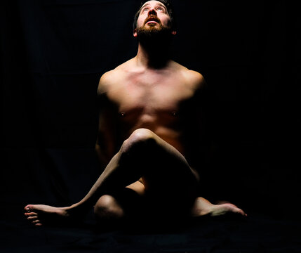 artistic male posing nude accenting muscular definition with crossed legs and overhead light with black background