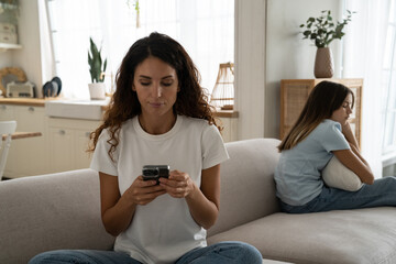 Adult Caucasian woman with mobile phone in hand sits on sofa on other side of depressed daughter....