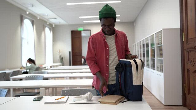 Never stop learning. Young African hipster guy student wearing spectacles putting textbooks into backpack after studying in university library, black man nerd preparing for exams. Higher education