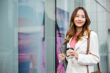 Young business woman wearing white suit jacket and smile while go to work outdoor mirror building background. Confident Businesswoman with cup of coffee holding Laptop walking outside office building