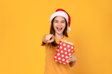Young woman wearing Christmas hat with holding a red bucket of popcorns while pointing front on blue background.