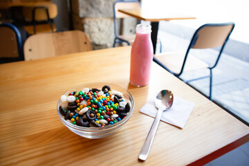 Dry breakfast in glass round plate bowl on a wooden table in cafe. Delicious cornflakes and pink milk in a bottle. Useful healthy breakfast. Food for kids. Different type cereals, multicolored balls.