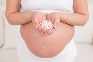 A faceless pregnant woman is holding a handful of white pills.