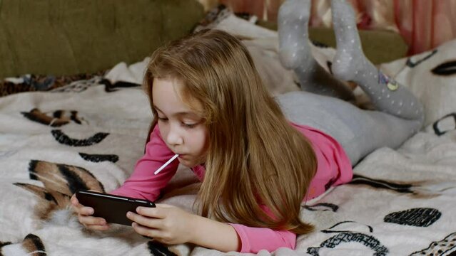 Cute Blonde-haired Girl With A Lollipop In Her Mouth, Shakes Her Legs, Enthusiastically Plays By Clicking On Smartphone Screen, Lying On A Sofa Covered With A Light Blanket. Digital Childhood Concept