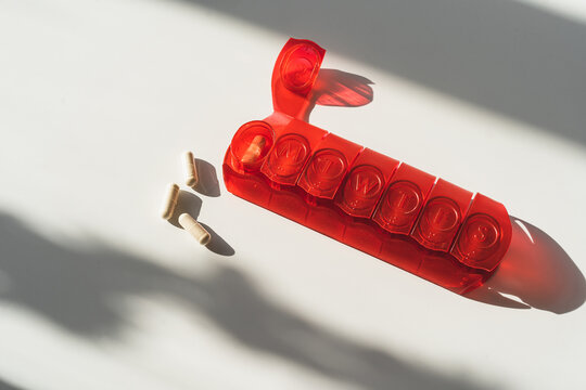 Closeup of red medical pill box with doses of tablets for daily take a medicine with white vitamins and capsules. Organizer for vitamins. Concept of health