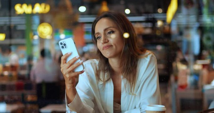 View through window glass of a cafe at a charming young woman relaxing, using smartphone, browsing websites, scrolling news feed, checking social media content, chatting with friends on the Internet