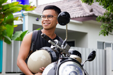 Smiling Filipino man with eyeglasses sitting on scooter holding vintage helmet. Male motorbike...