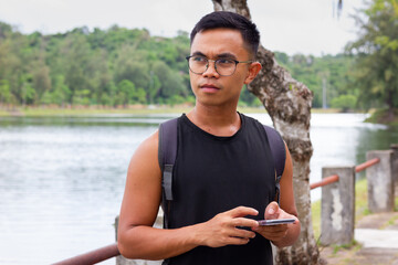Fototapeta premium Serious Filipino man with eyeglasses holding cellphone with lake on background. Asian male thinking with phone in hands outdoors