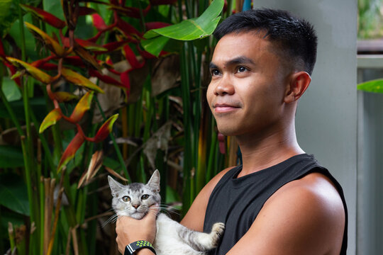Smiling Filipino young man holding gray cat at home backyard. Male owner petting kitten in his arms. Animal love, pet adoption concepts