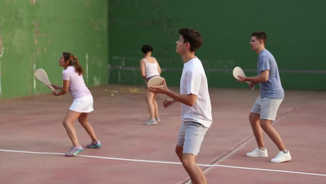 Group of people playing pelota at open-air fronton in summer, swinging traditional wooden bat to return ball. Sportsman ready to hit volley