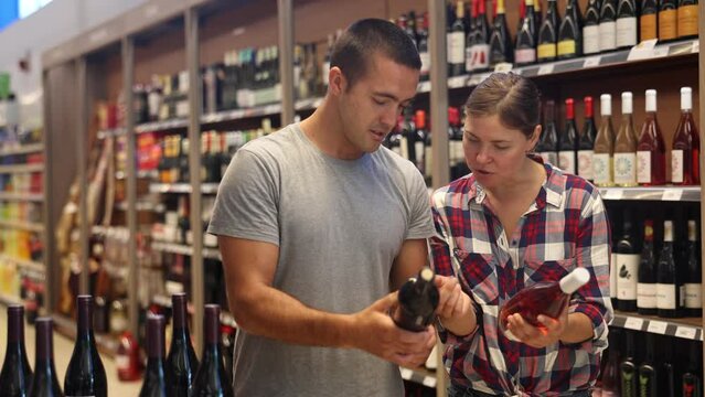 Positive young adult couple looking for perfect wine for solemn occasion in supermarket wine department. High quality 4k footage