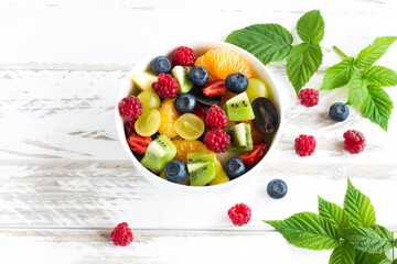 Fototapeta na wymiar fruit salad with banana, kiwi, grapes, raspberries, blueberries in a bowl on a white wooden table.