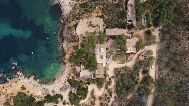 Aerial top view of abandoned hotel Sert at Cala d&rsquo;en Serra Beach, Spain and clear blue ocean with rock cliffs, roads and trees.