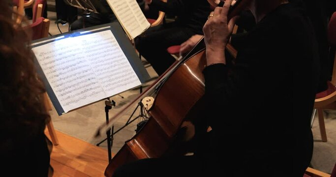 Medium handheld shot of a talented violoncello player during a concert with orchestra in theatre to classical music by mozart with view of music stand with sheet music and other musicians. Slow motion