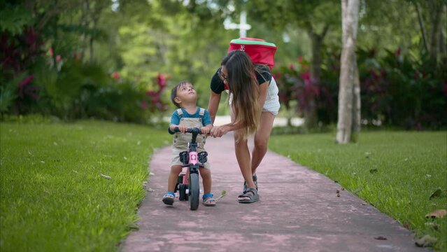 Latin baby boy riding a pink training bicycle on the road at the park being helped by his mom