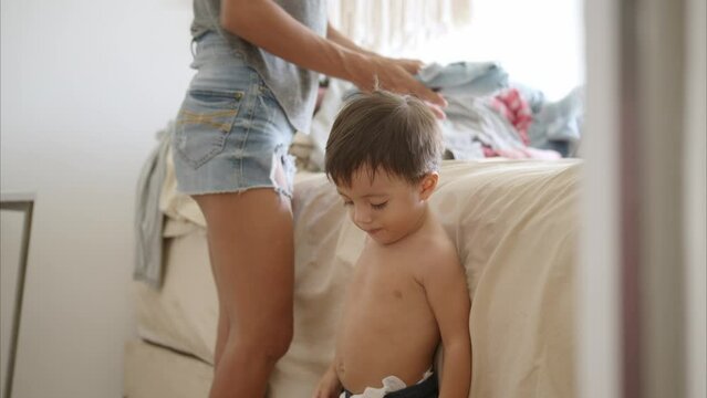 Little latin shirtless baby boy in blue shorts helping his mother fold clothes on the beth at their bedroom