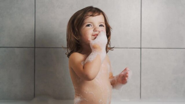 A cute two-year-old girl washes herself with foam in the bath