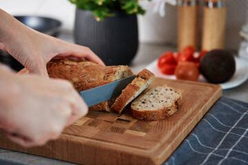 Woman cutting loaf of whole grain bread with large knife on cutting board. Hands slicing fresh...