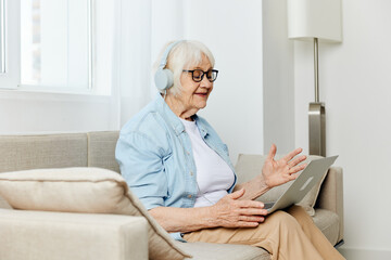 a happy, emotional elderly woman with gray hair is sitting at home on a cozy sofa with headphones on her head and happily smiling holding a laptop on her lap