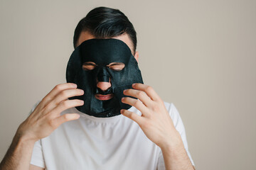 Close-up portrait adult man applying a black moisturizing paper mask isolated over gray background. Beauty treatment. Facial skincare.