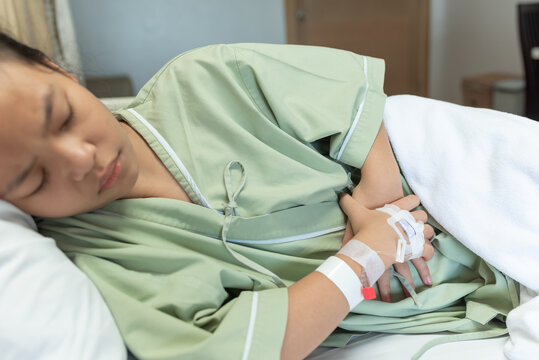 Asian young beautiful female lying on the bad having stomach ache and IV saline drip to back of hand lying on bed in hospital, medical and health concepts.