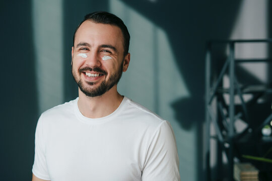 Close up portrait of smiling satisfied handsome enjoying skincare procedure after shower, standing at home. Facial treatment, cosmetology, beauty concept