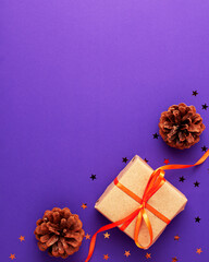 Flat lay of brown gift box with a orange satin ribbon bow and stars shapes pine cone on vibrant purple background at the bottom with copy space. Holiday autumn and Halloween concept