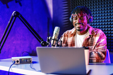African radio host sitting at desk recording in studio with microphone and laptop