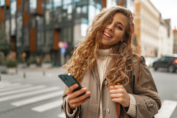Obraz premium Smiling curly woman wearing warm coat walking down the street and using her phone