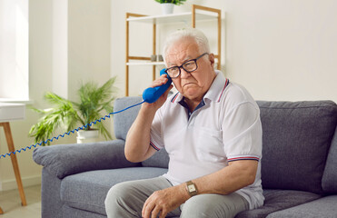 Elderly man incorrectly holds landline telephone handset to his ear and does not understand why he cannot hear anything. Confused distracted pensioner sitting on sofa at home and listening to landline - obrazy, fototapety, plakaty
