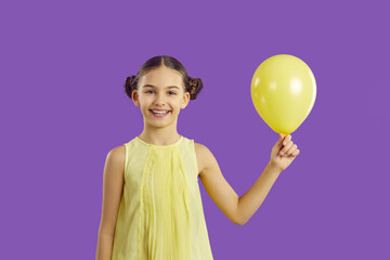 Happy pretty child with yellow balloon. Cheerful little girl with cute hair buns isolated on purple background holding mock up balloon, looking at camera and smiling. Kids, fun, birthday party concept