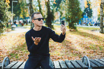 A lonely young man in a black sweater sits in the park on a bench and talks on the phone. A warm autumn day