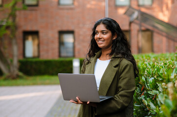 Brunette indian woman smiling while working with laptop on city street